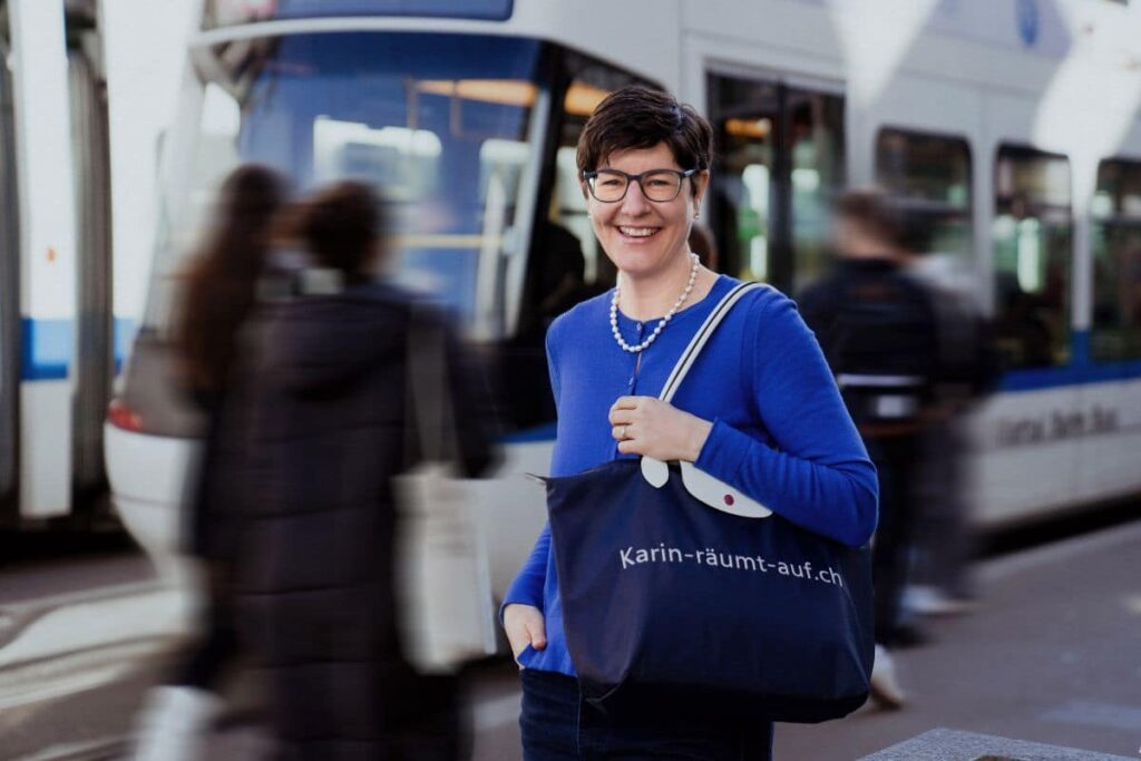 Ordnungscoach Karin Treichler steht vor einem ankommenden Tram und trägt ihre Tasche mit der Aufschrift Karin-räumt-auf.ch